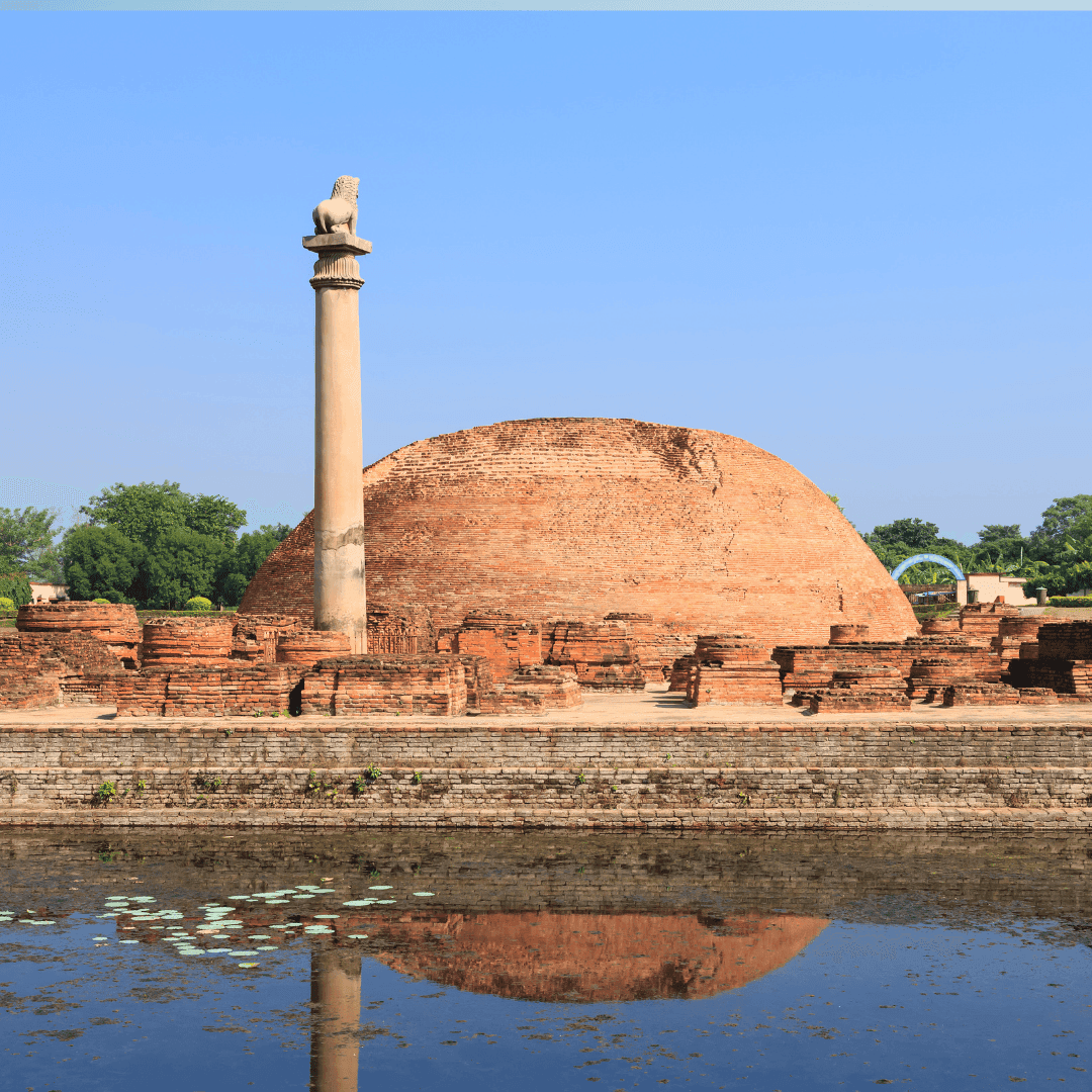 "The ancient sandstone Ashokan Pillar at Vaishali, topped with a single seated lion capital, standing before a brick stupa under a clear sky."